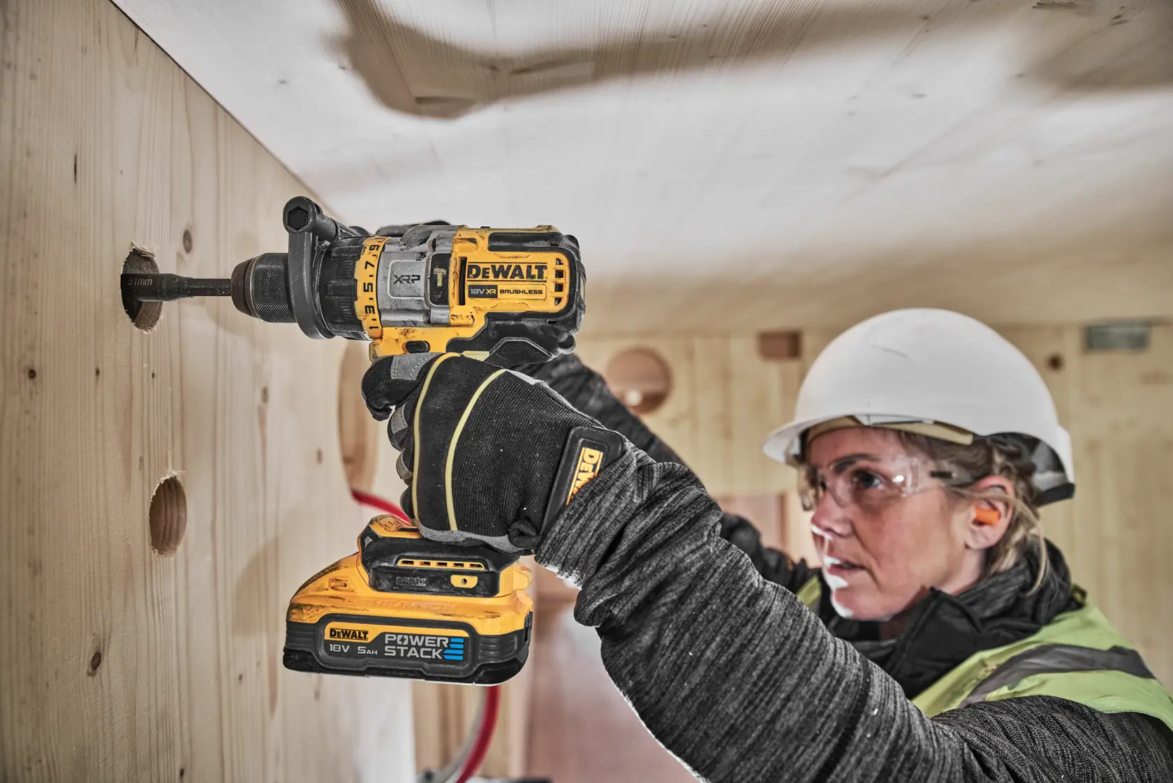 En person med verneutstyr bruker en DEWALT DCD999H2T trådløs drill for å bore et hull i en trevegg. Drillingen har et POWERSTACK-batteri.