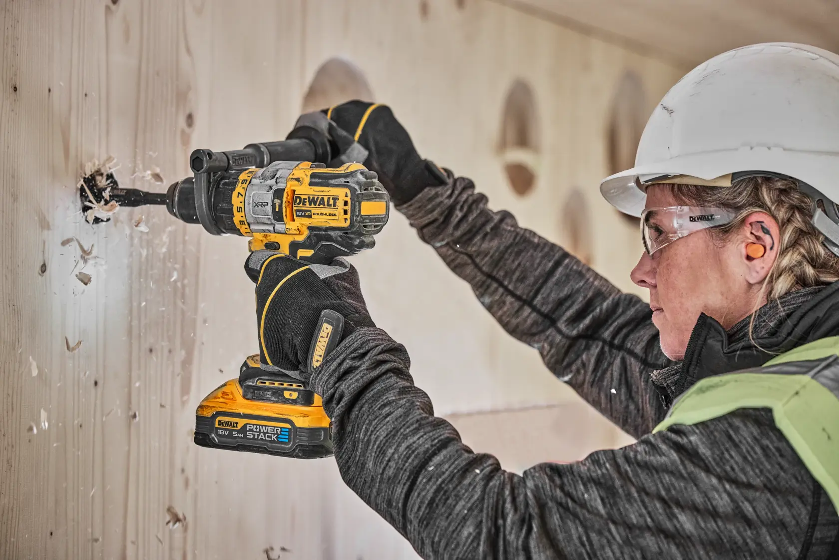 En person med sikkerhetsutstyr bruker en DEWALT DCD999H2T batteridrill for å bore et hull i en trevegg.