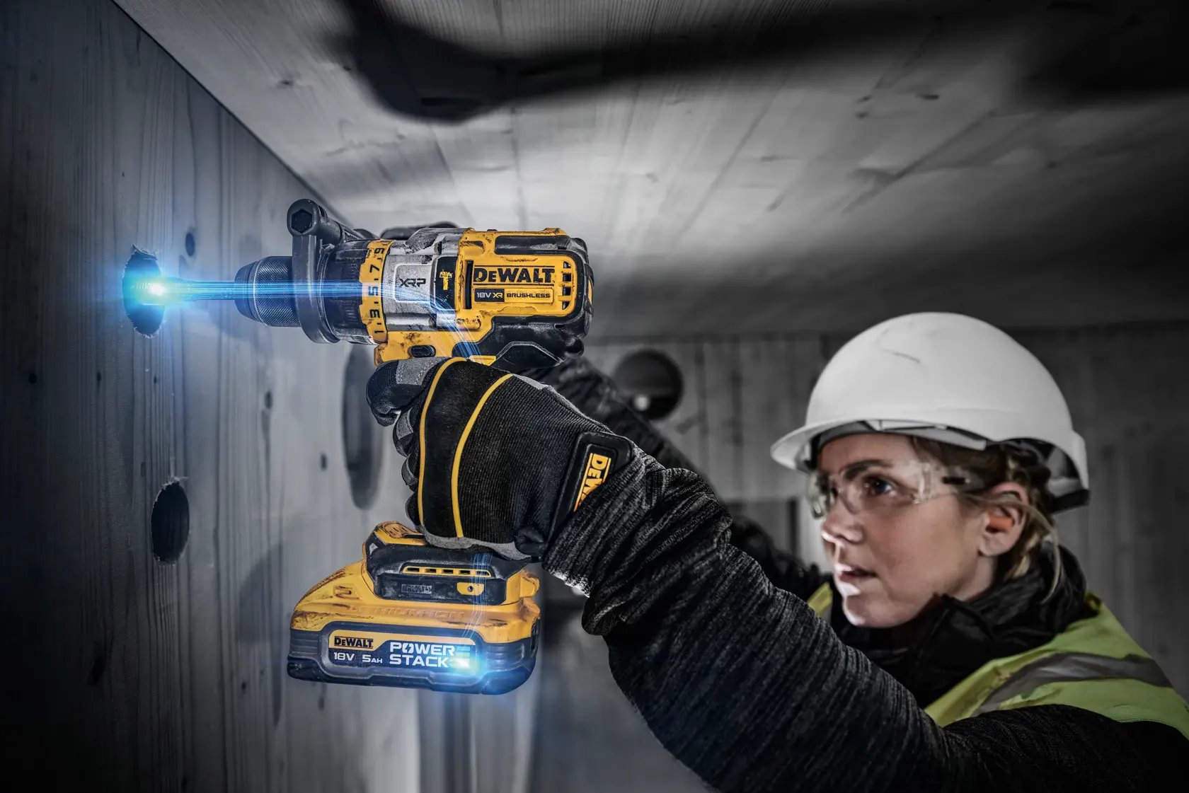 En person med verneutstyr bruker en DEWALT DCD999H2T PowerStack batteridrill for å bore et hull i en vegg av tre. Drillens lys er på, og personens ansikt er skjult for personvern.