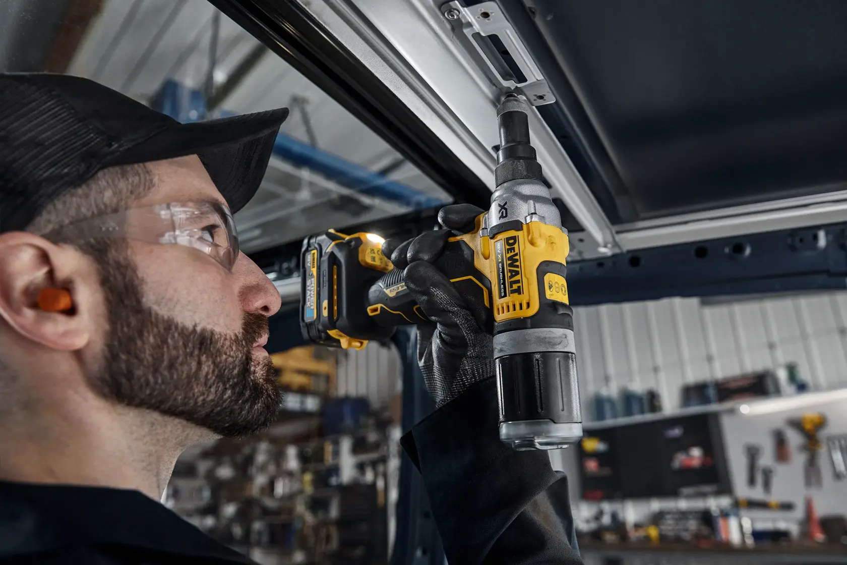 En person med hørselsvern og hansker bruker et DEWALT DCF414 batteriverktøy for å feste eller bore i en metallramme, med et verksted i bakgrunnen.