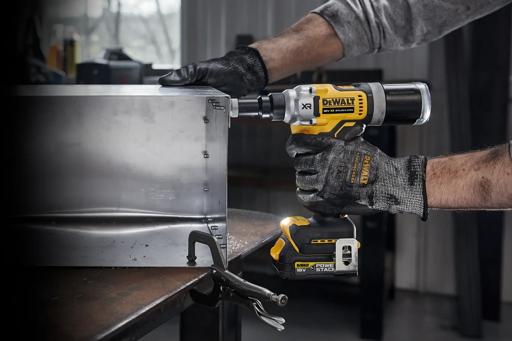 En person med hansker bruker et DEWALT 18V XR batteridrevet popnagleverktøy (modell DCF414) for å feste metallplater på et arbeidsbord i et verksted.