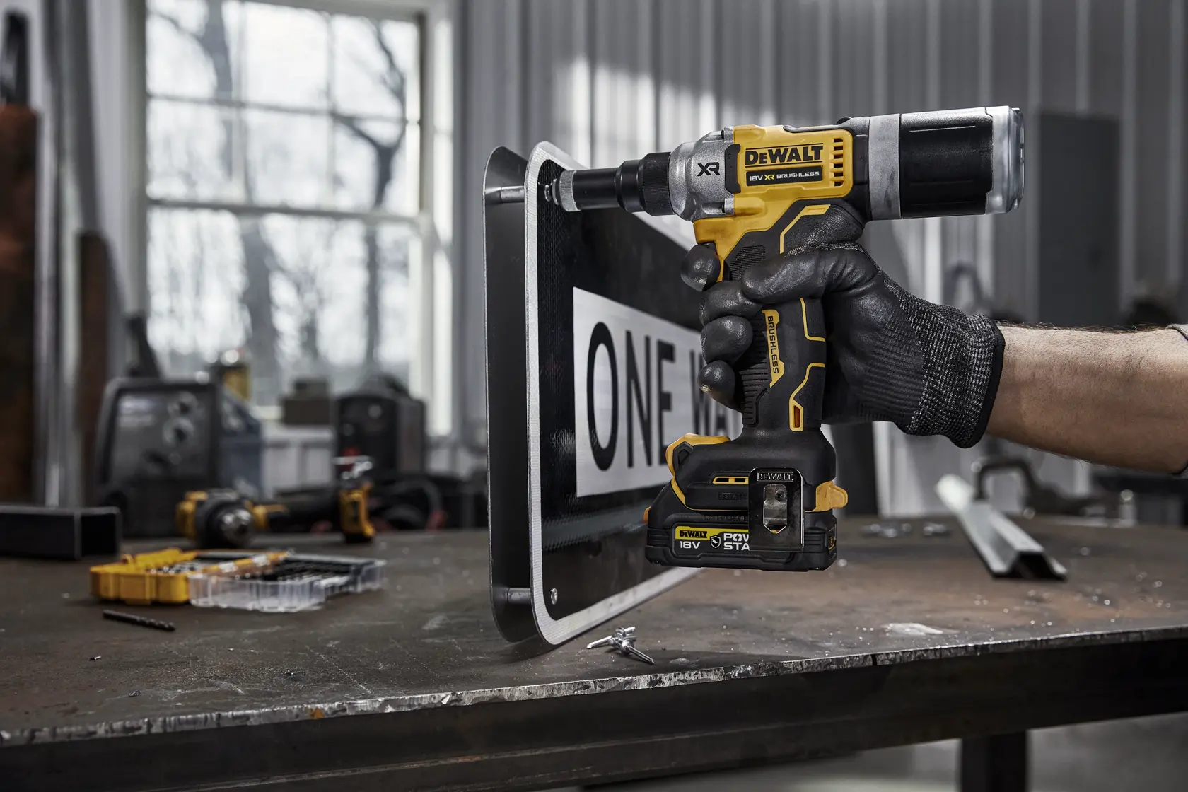 En person med svart hanske bruker et gul-svart DEWALT DCF414E2GT elektroverktøy for å bore eller feste et gateskilt på en metallarbeidsbenk i et industriverksted.