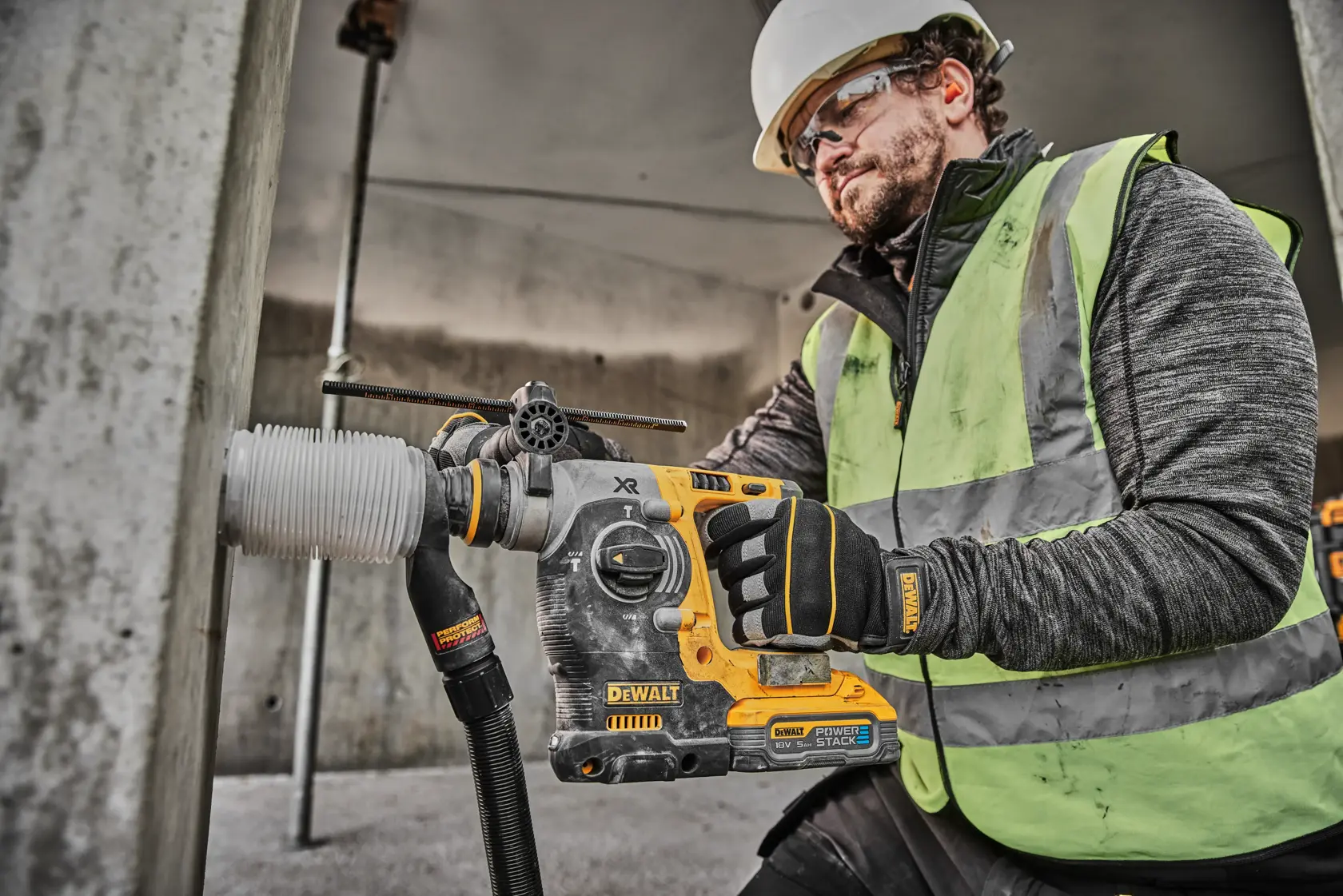 En bygningsarbeider med hjelm og synlighetsvest bruker en DEWALT DCH273H2T batteridrill for å bore i en betongsøyle. Arbeideren har også DEWALT-hansker.