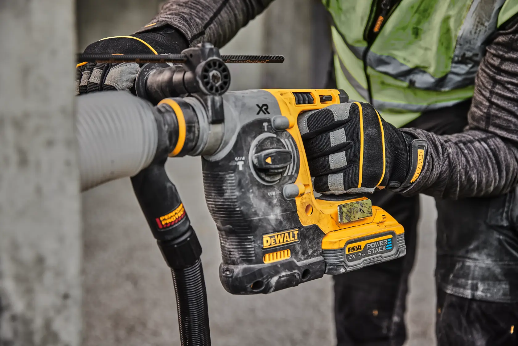 Nærbilde av en arbeider som bruker en DEWALT batteridrevet rotasjonshammer med POWERSTACK-batteri for å bore i betong, iført vernehansker og klær.