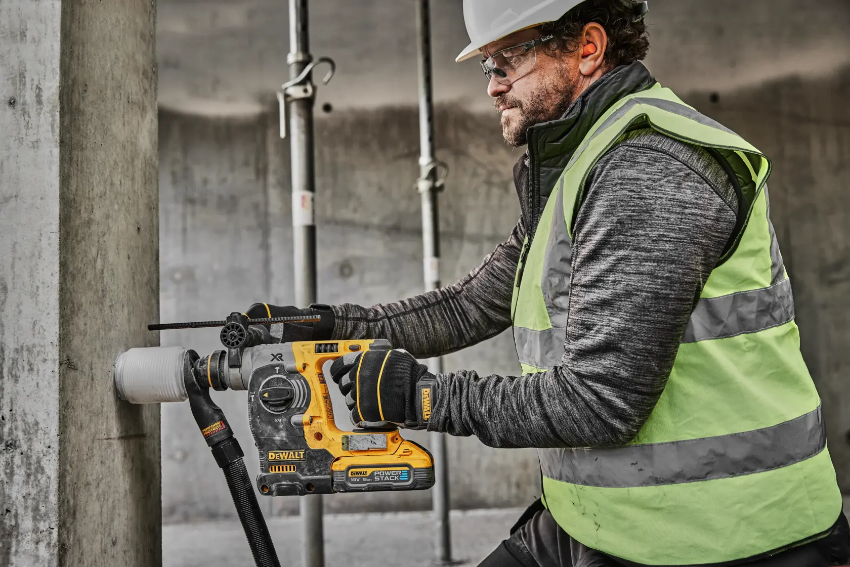 En person med sikkerhetsvest og hansker bruker en DEWALT DCH273H2T trådløs borhammer til å bore i en betongsøyle på en byggeplass. Brukerens ansikt er utvisket av hensyn til personvern.