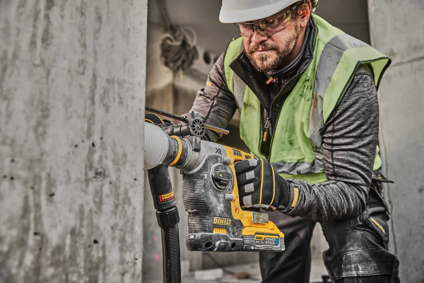 En anleggsarbeider bruker en DEWALT DCH273H2T roterende hammerboremaskin med støvavsug, og borer i betong. Arbeideren har på seg sikkerhetshansker, refleksvest og hjelm. Boremaskinen er fra DEWALT og har PowerStack-batteri.