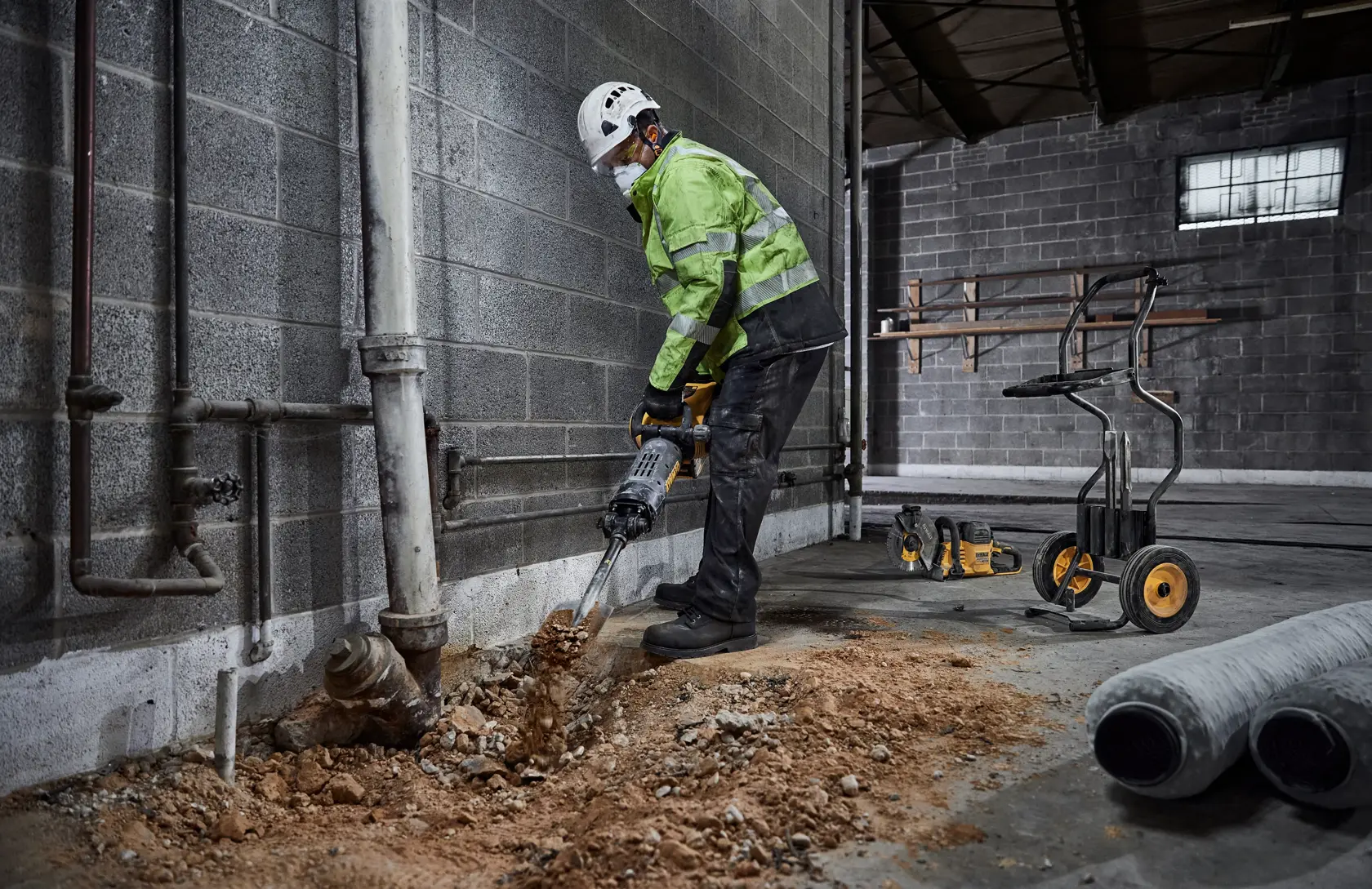 En person med verneutstyr bruker en DEWALT rivningshammer for å bryte opp et betonggulv nær en vegg i et industrielt miljø. Byggeutstyr og verktøy er synlig i bakgrunnen.