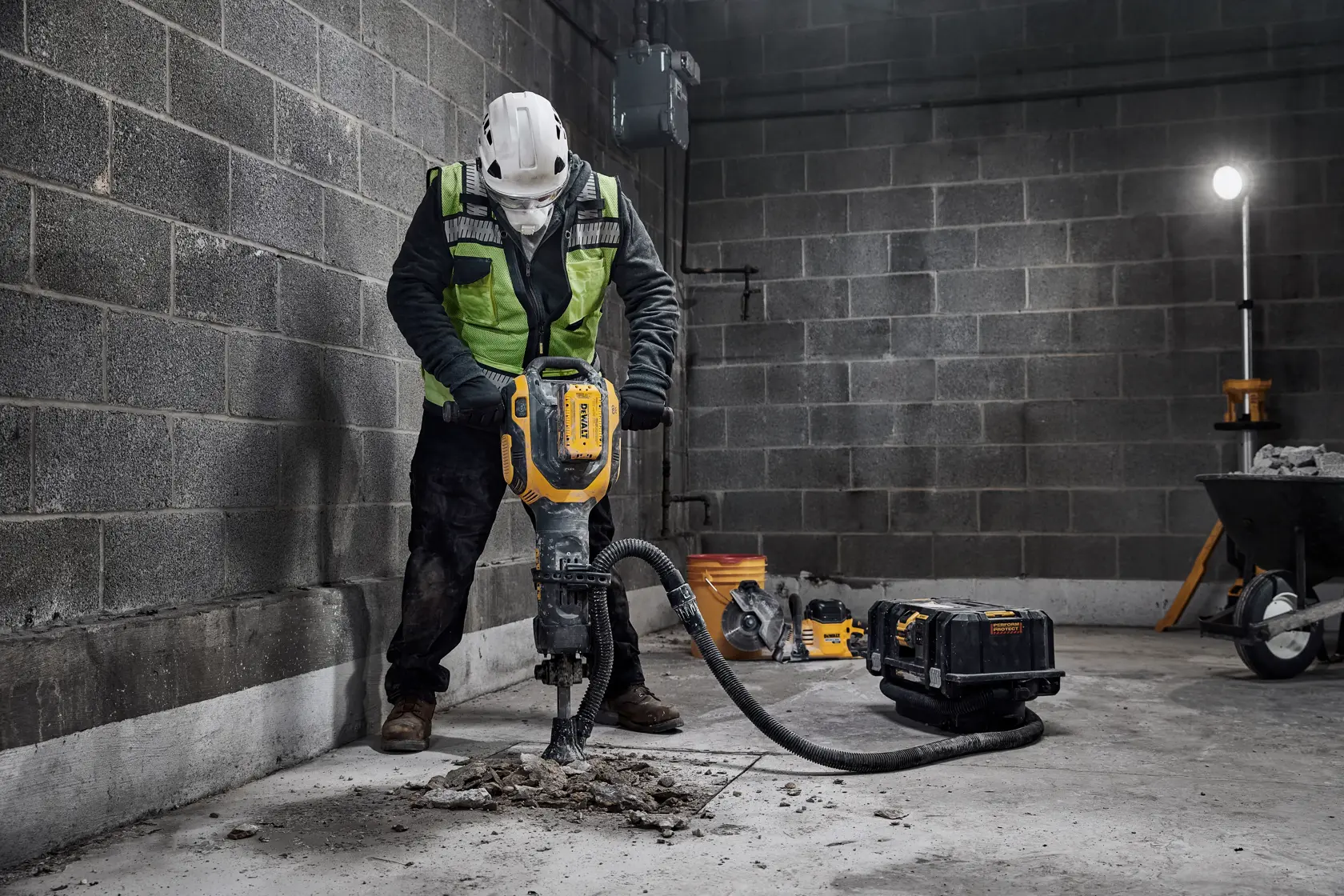 En bygningsarbeider i verneutstyr bruker en elektrisk DEWALT-pigghammer for å bryte opp betong på en byggeplass. Verktøyet er koblet til et støvavsugssystem, og det er byggeutstyr i bakgrunnen.