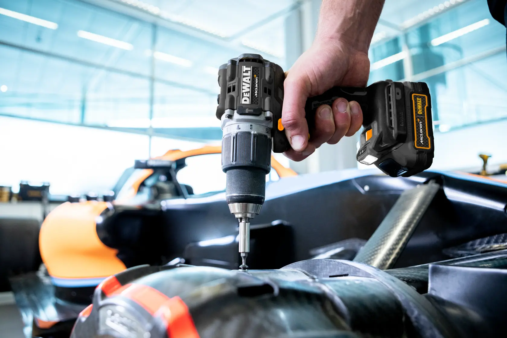 Nærbilde av en person som bruker en DEWALT batteridrevet drill for å feste eller montere deler på det som ser ut til å være en kjøretøykomponent, med en oransje bil synlig i bakgrunnen.