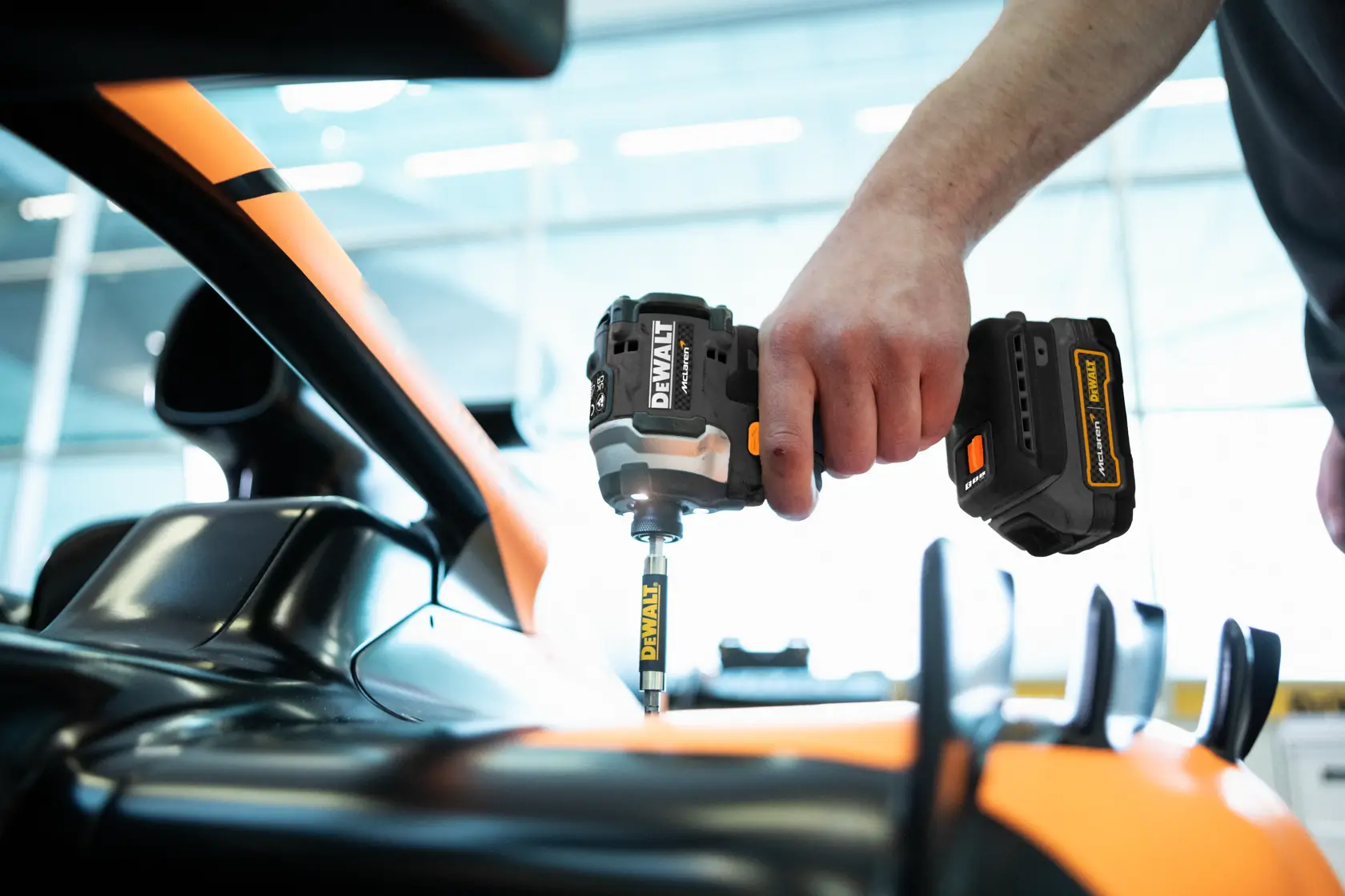En person bruker en DEWALT batteridrevet slagtrekker for å arbeide på et svart og oransje kjøretøy, sannsynligvis for vedlikehold eller montering i et godt belyst innemiljø.