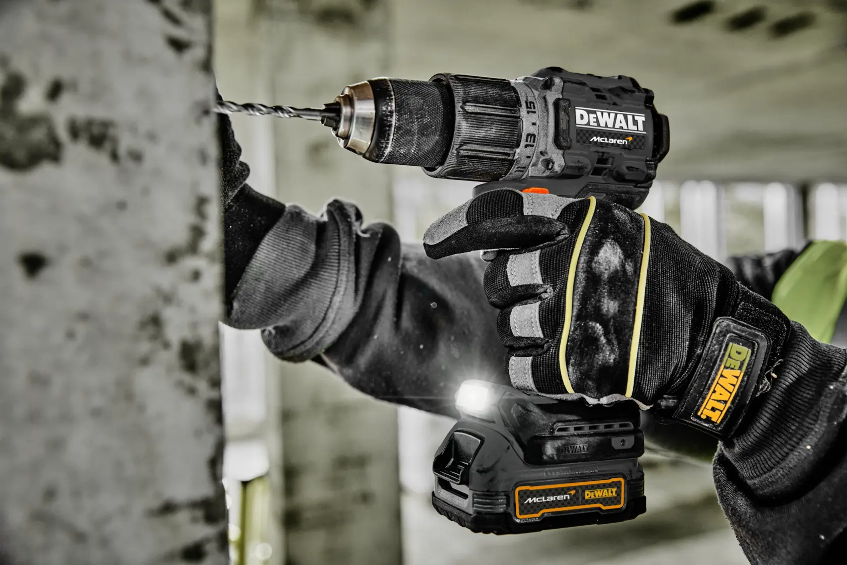 En person med vernehansker bruker en DEWALT batteridrill til å bore i en betongvegg. Drillens batteripakke er fra McLaren og har LED-lys. Scenen er på en byggeplass.