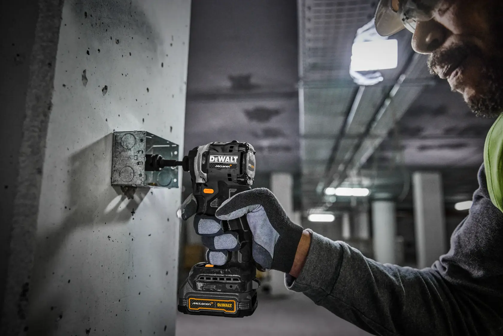 En person med sikkerhetshansker bruker en trådløs DEWALT drill for å feste en metallbeslag til en betongvegg i et industrielt miljø.