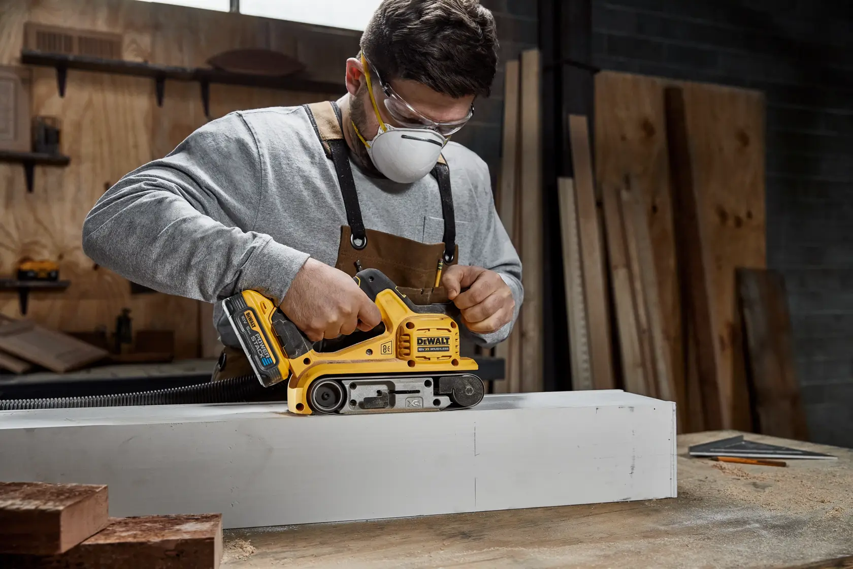 En person med forkle bruker en gul DEWALT DCW220N batteridrevet båndsliper for å slipe et stort trestykke i et verksted.