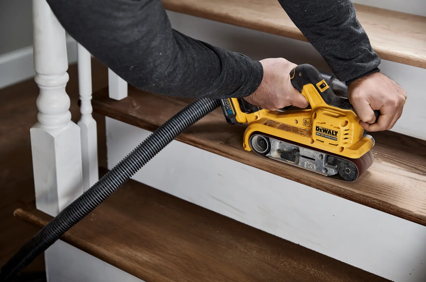 En person bruker en DEWALT DCW220N trådløs båndsliper for å slipe tretrapper, med en støvsugerslange festet til verktøyet for oppsamling av støv.