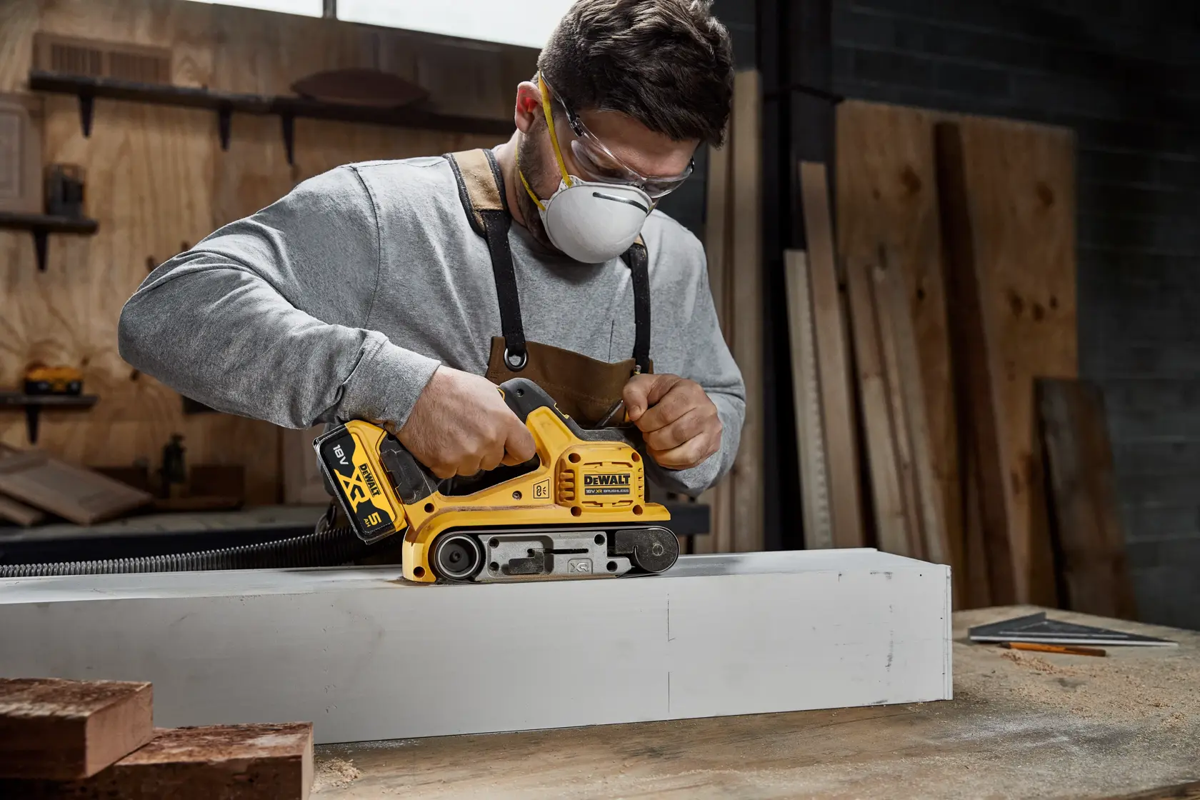 En person bruker en DEWALT batteridrevet båndslipemaskin (SKU: DCW220P2) på et trestykke i et verksted. Personen har på seg vernebriller og forkle, og ansiktet er uskarpt.