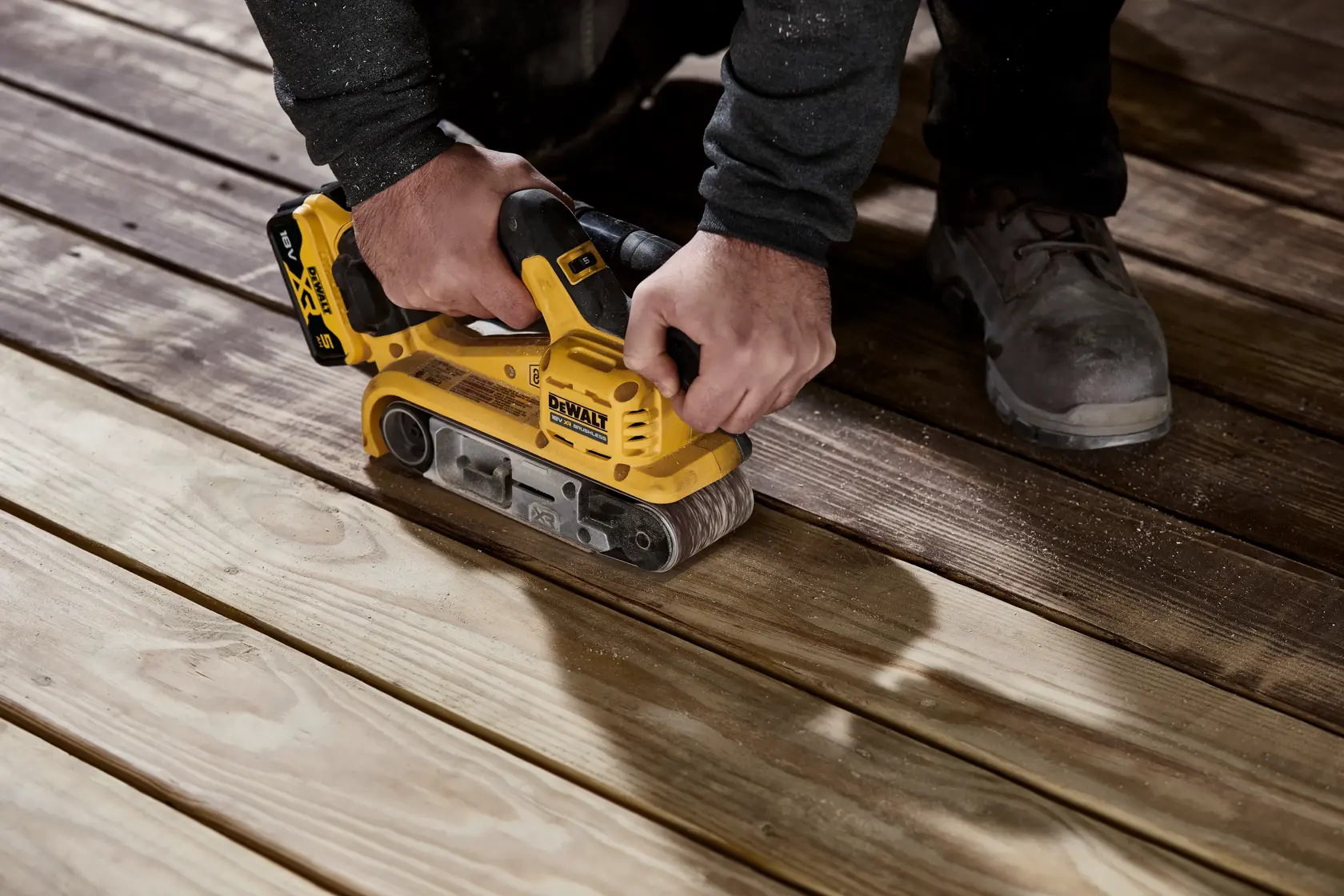 En person bruker en gul DEWALT batteridrevet båndsliper (modell DCW220P2) til å slipe tregulvplanker.