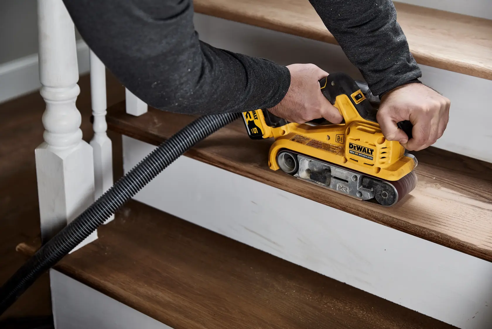 En person bruker en gul DEWALT DCW220P2 trådløs beltessliper til å slipe et trappetrinn i tre. En støvsugerslange er koblet til verktøyet for støvoppsamling.