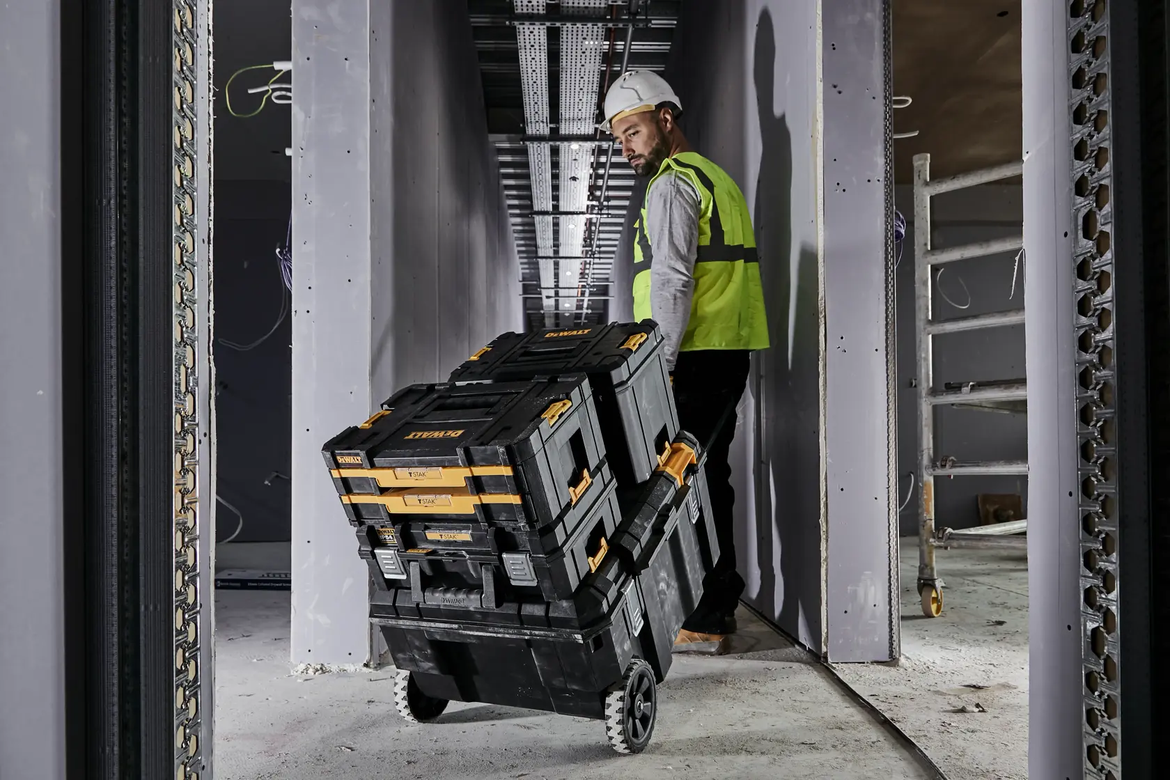 Worker pulls stacked DEWALT toolboxes on wheels at construction site in a narrow hallway.