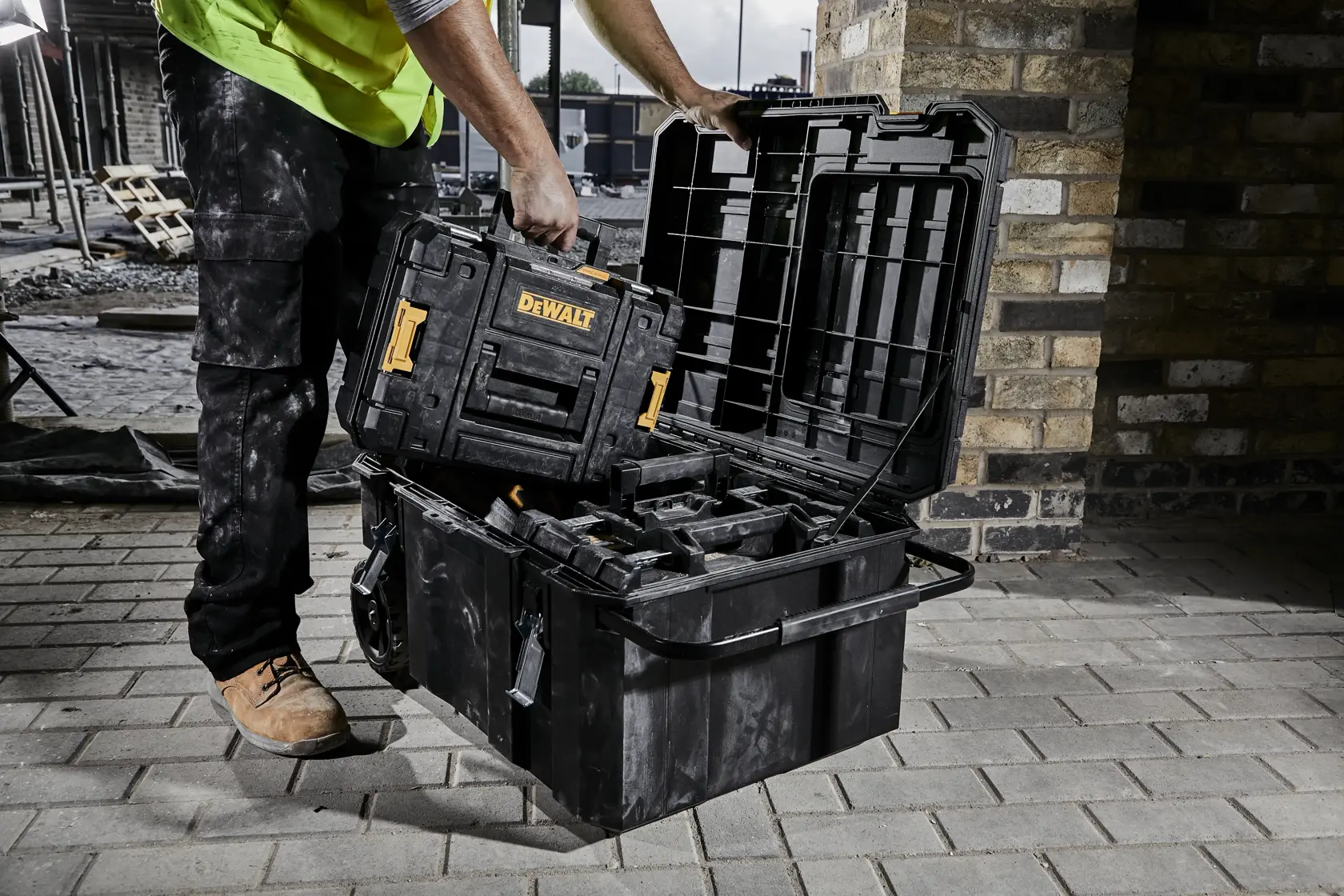 Person handles DEWALT toolbox, placing it in a large open storage case on a construction site with brick wall.