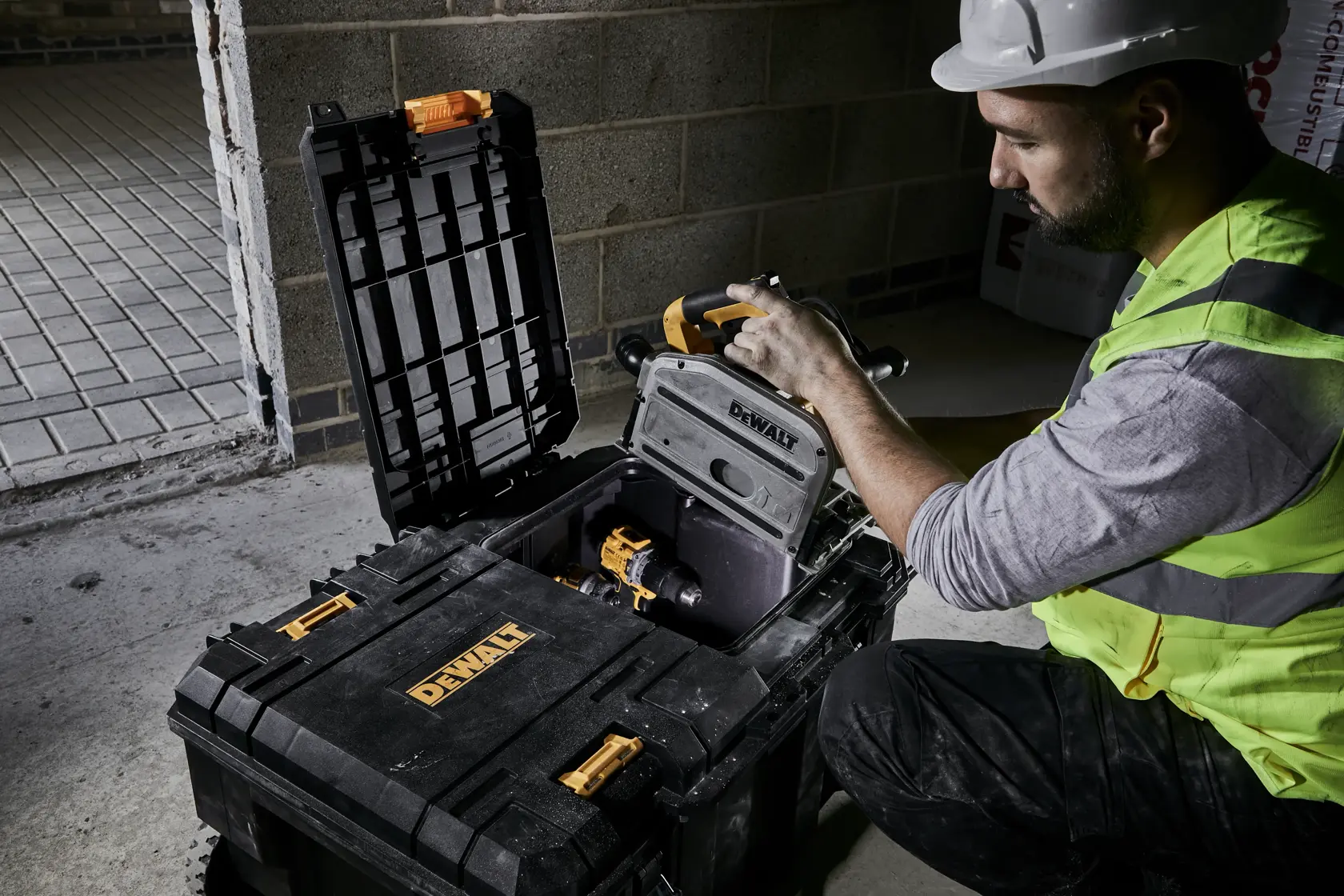 Worker opening a DEWALT toolbox with power tools inside, on a construction site with a concrete wall.